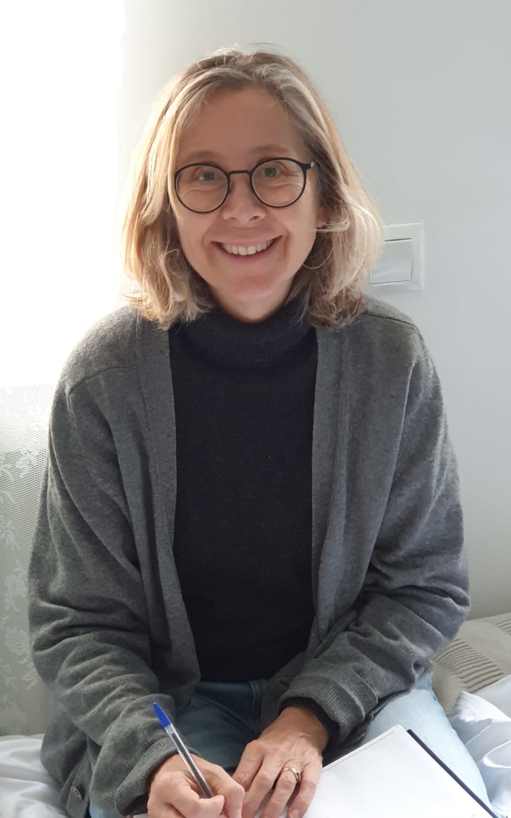 A photograph of Ruth smiling. She has long blonde hair, round glasses, and a grey cardigan.