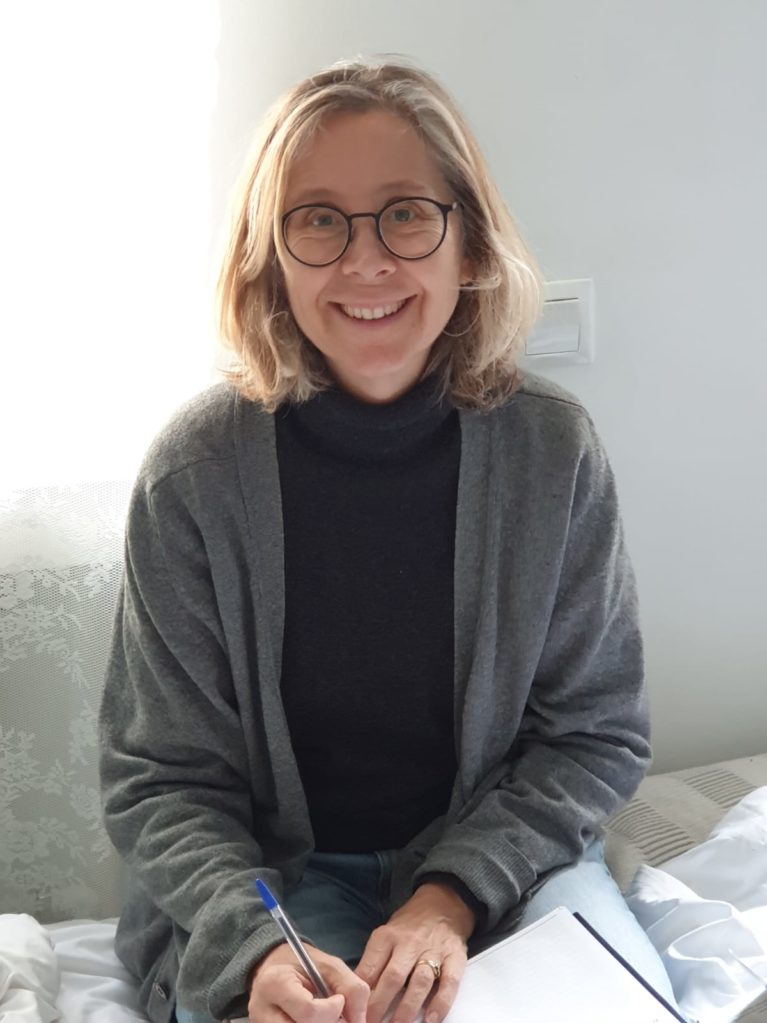 A photograph of Ruth smiling. She has long blonde hair, round glasses, and a grey cardigan.